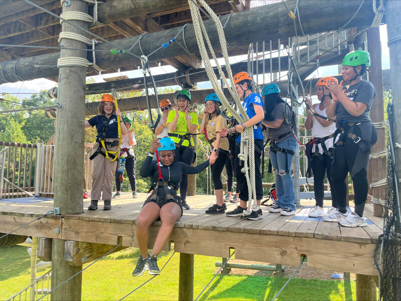 A group of people are on a ropes course, wearing helmets and harnesses. They appear to be navigating various obstacles and platforms. The course is set up in a wooded area, with trees visible in the background. The participants seem to be enjoying the activity, with some smiling and laughing.